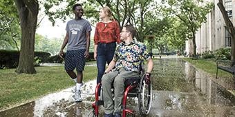 Three students, one in a wheelchair, pass close to the Catehdral of Learning together while talking on a rainy day.